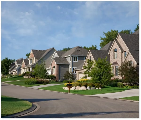A suburban neighborhood street lined with large, modern single-family homes featuring well-maintained lawns, colorful flower beds, and mature trees. The houses have a variety of architectural styles, including brick and siding exteriors, gabled roofs, and multiple windows. The street is quiet, curving gently with neatly edged sidewalks and driveways leading up to each house.